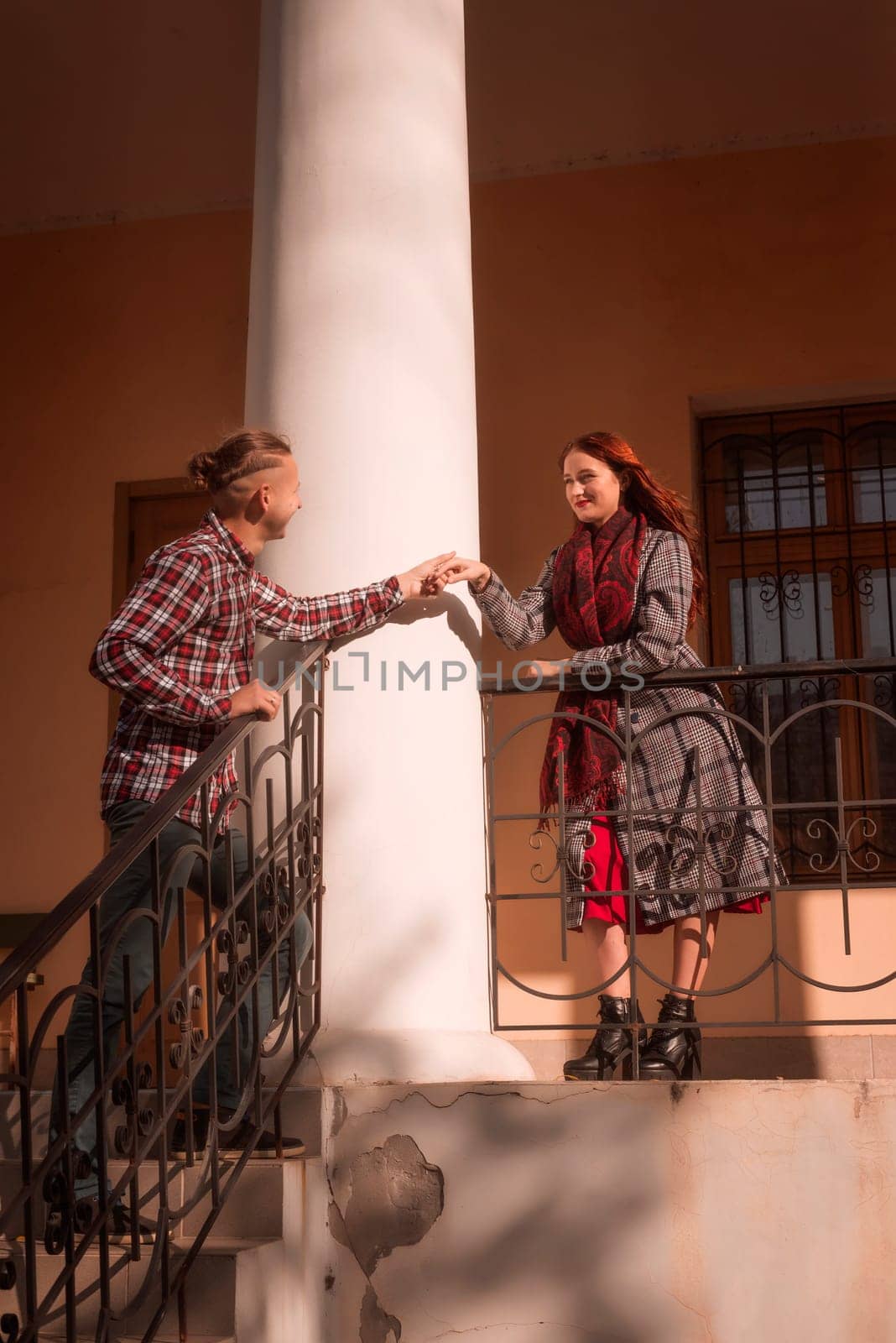 Young couple in love looking at each other romantically together. against the backdrop of an old building, in the rays of the sun. the girl is wearing a coat and a red scarf, with a hat.