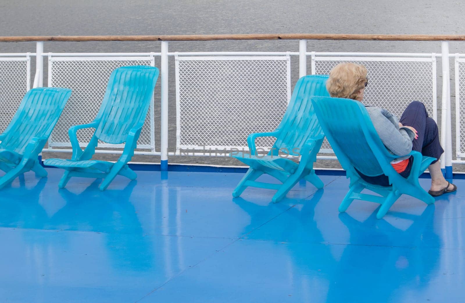 A lone middle-aged woman sits in a chaise lounge on the deck of a cruise ship, looking out to sea. Unrecognizable face.