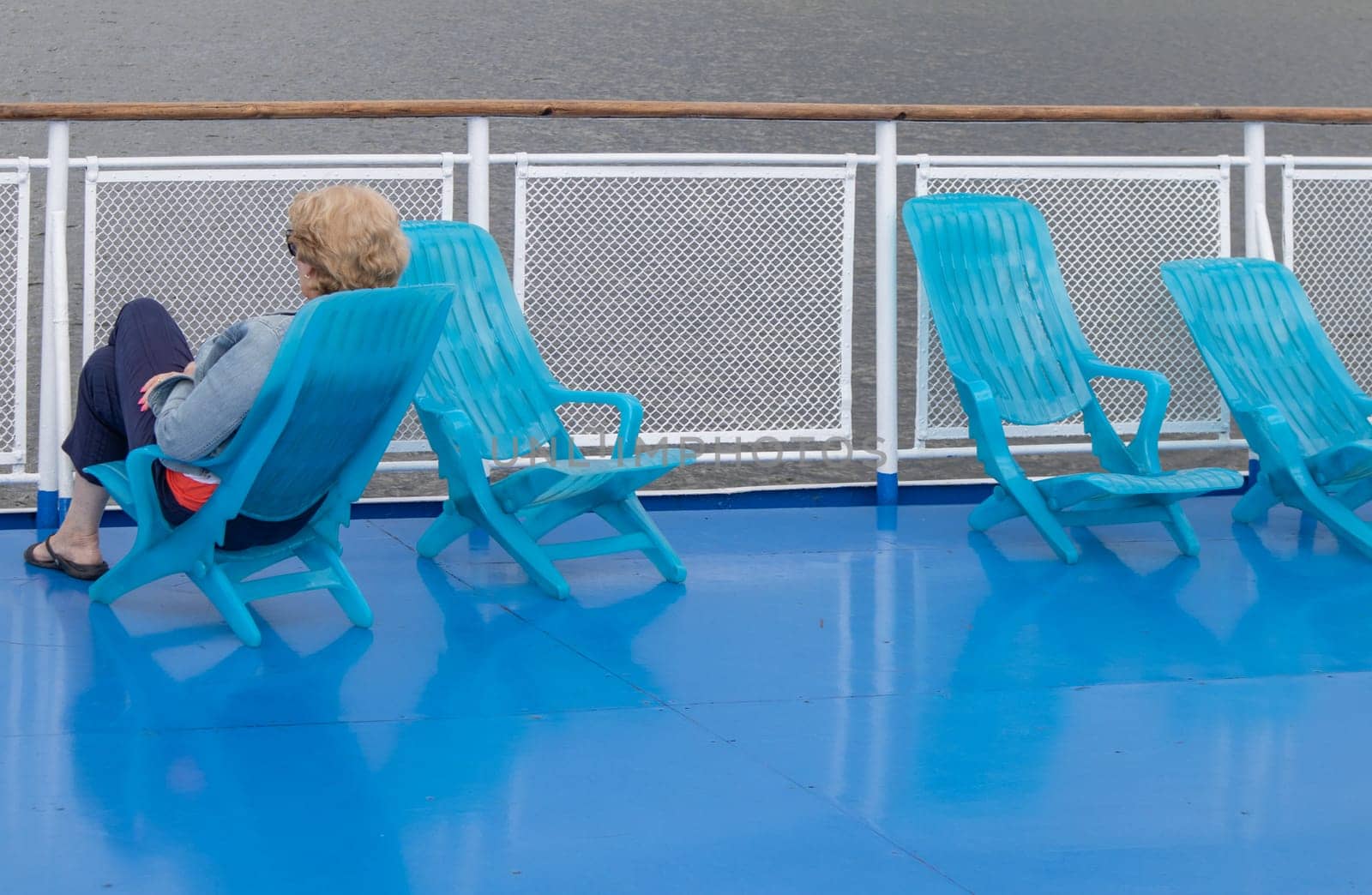 A lone middle-aged woman sits in a chaise lounge on the deck of a cruise ship, looking out to sea. Unrecognizable face.