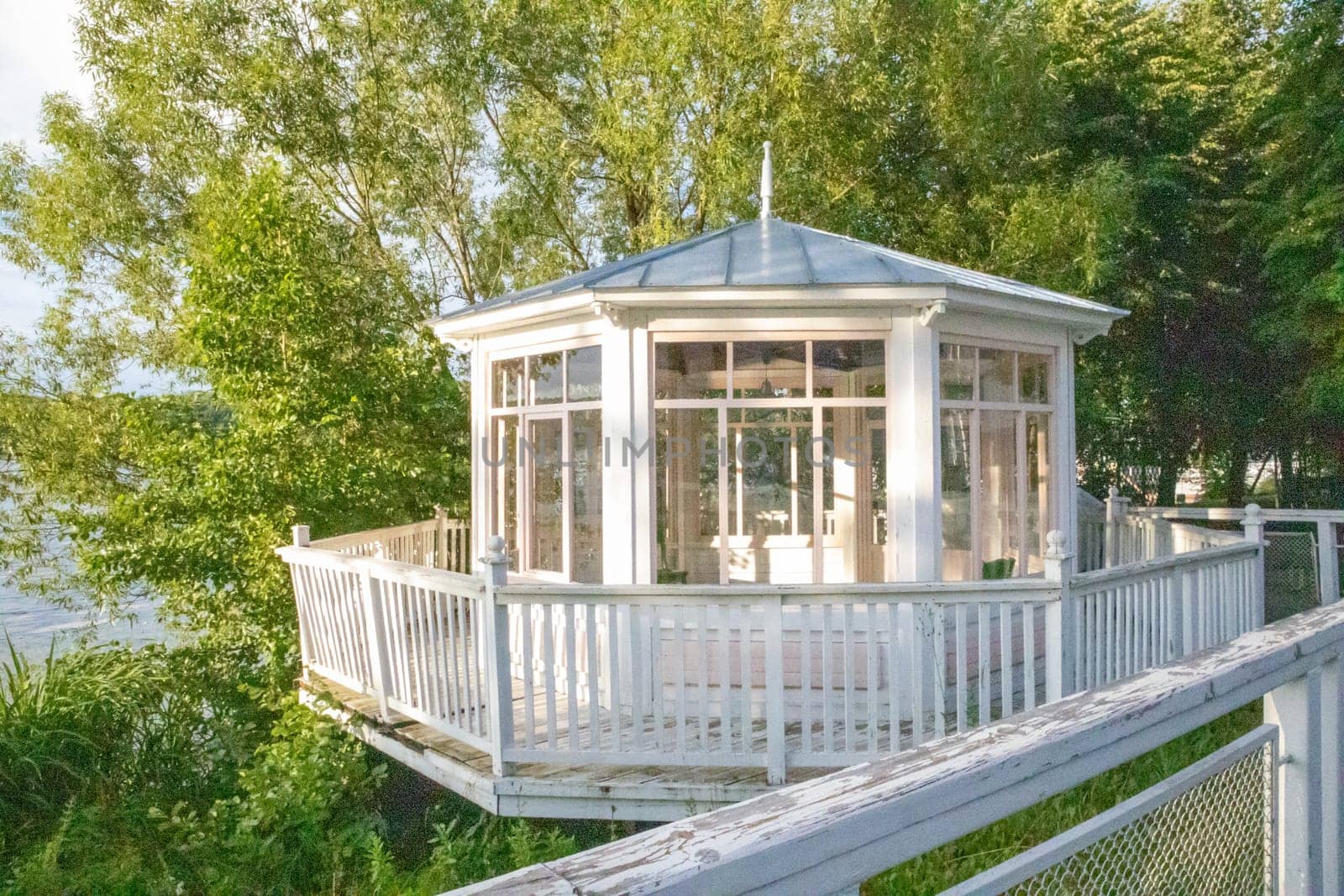 White wooden vintage gazebo with glass walls against a backdrop of green trees on a sunny summer day.