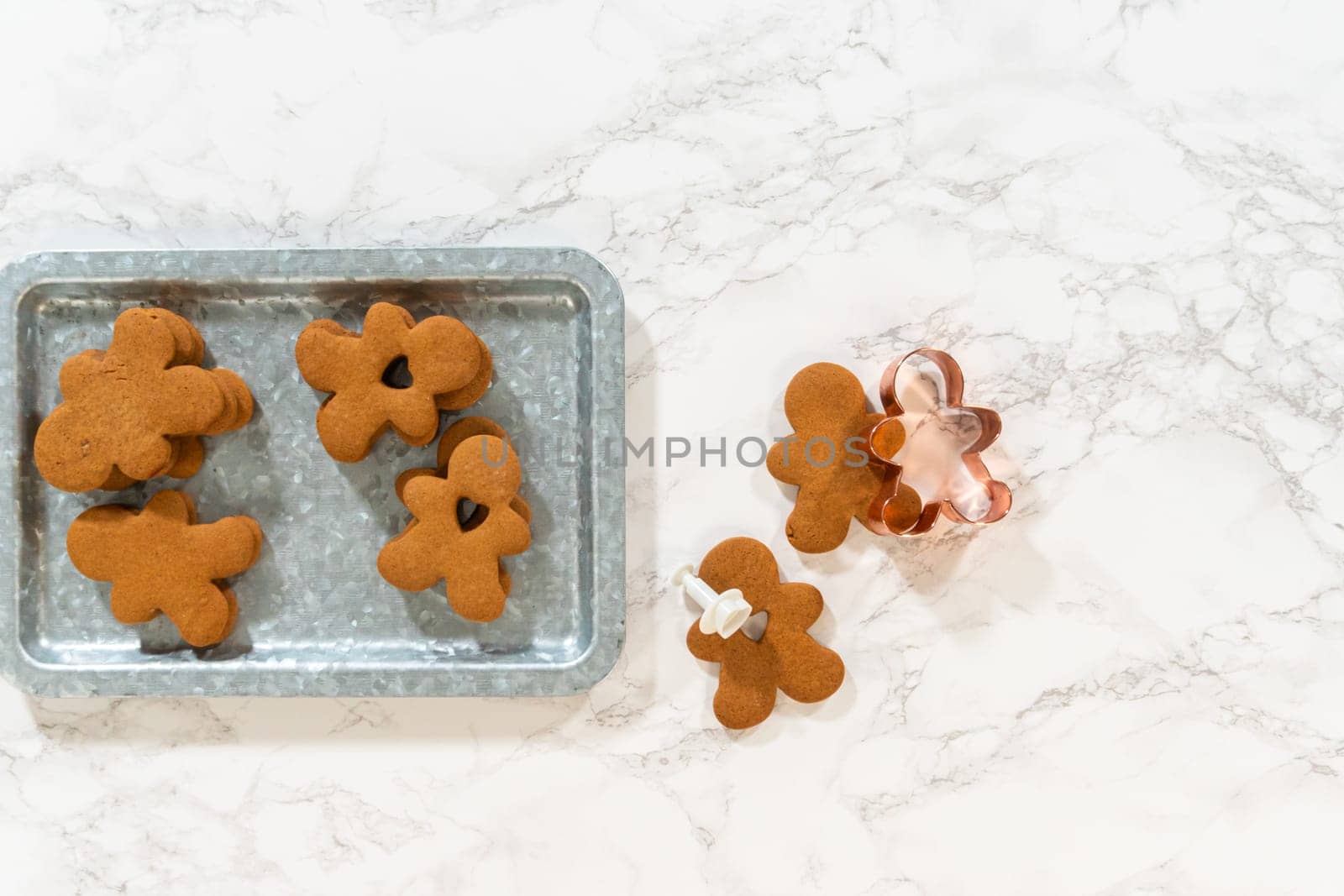 Flat lay. Gingerbread cookies, including a gingerbread man with a heart-shaped cutout, rest on a rustic metal tray against a marble countertop.