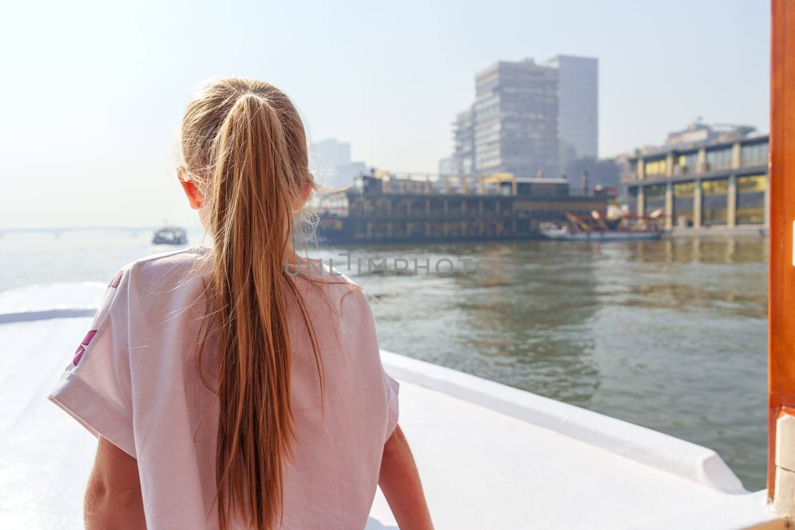 With her long hair flowing, a young girl gazes at the city skyline from a boat. The sun glints off the water, adding warmth to the vibrant urban atmosphere.