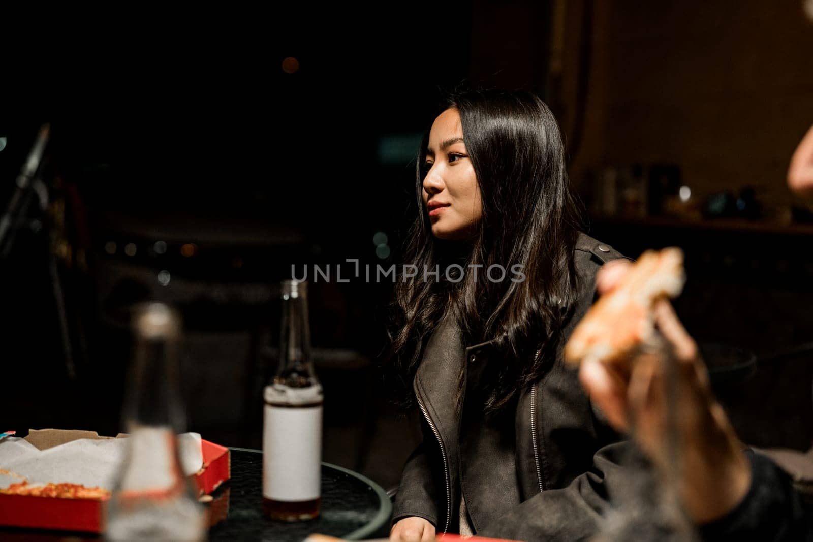A young woman with long black hair wears a leather jacket while having pizza with friends at a nighttime gathering. The atmosphere is relaxed and casual.