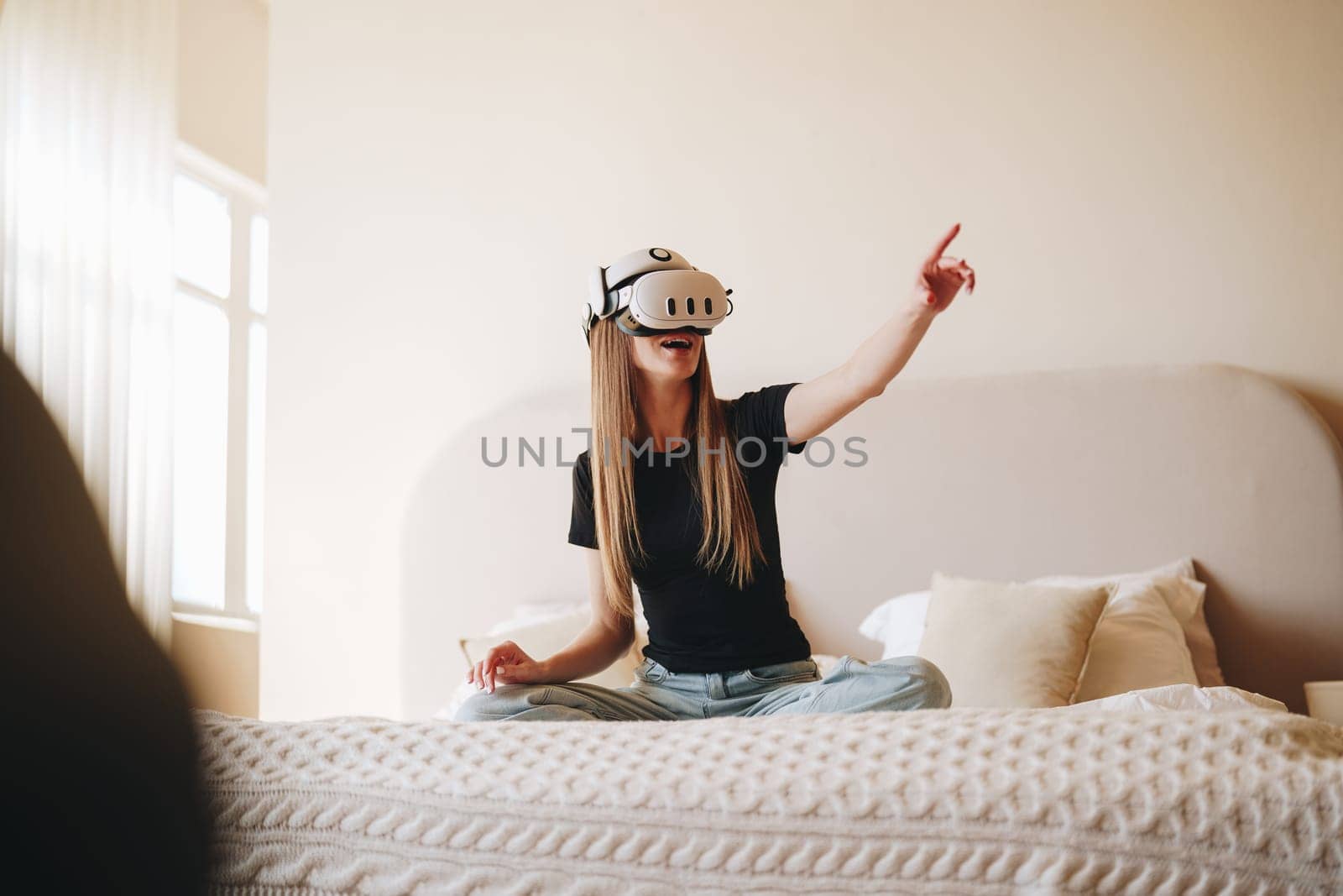 A young woman engages with a virtual reality headset while sitting cross-legged on a bed. Natural light fills the cozy room, creating an inviting atmosphere for her immersive adventure.