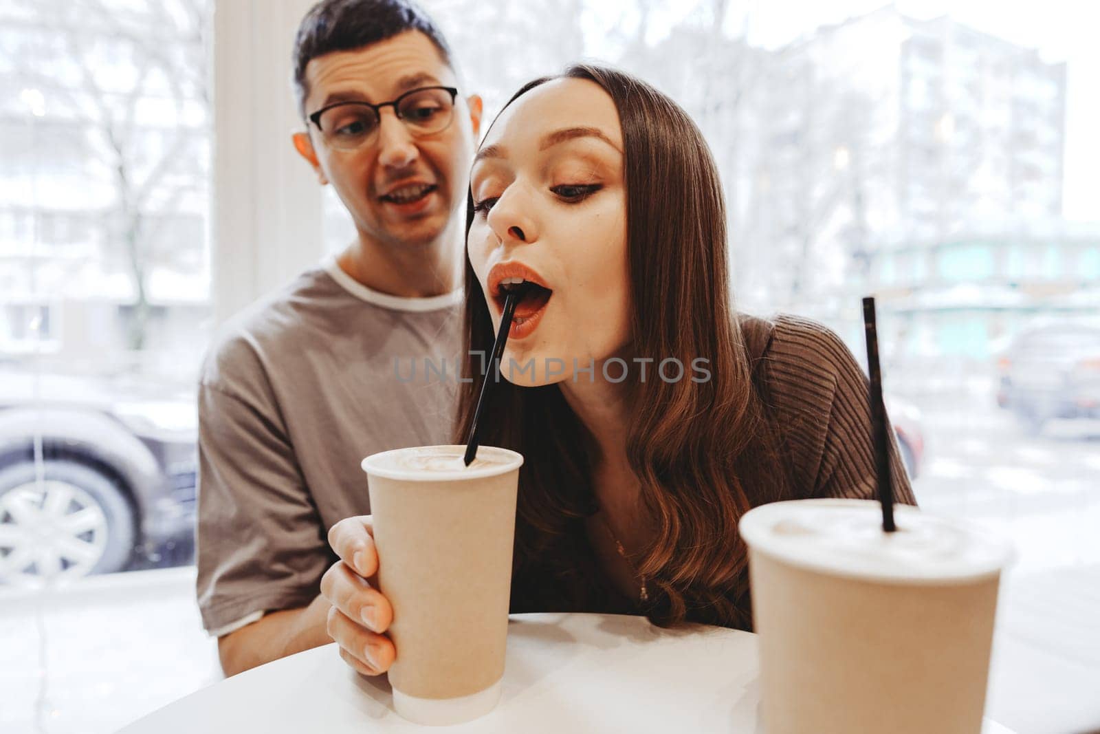 A young couple sits at a table in a warm cafe, smiling and sipping on their drinks. The rain falls outside, creating a cozy atmosphere. They are engaged in a fun and affectionate interaction.