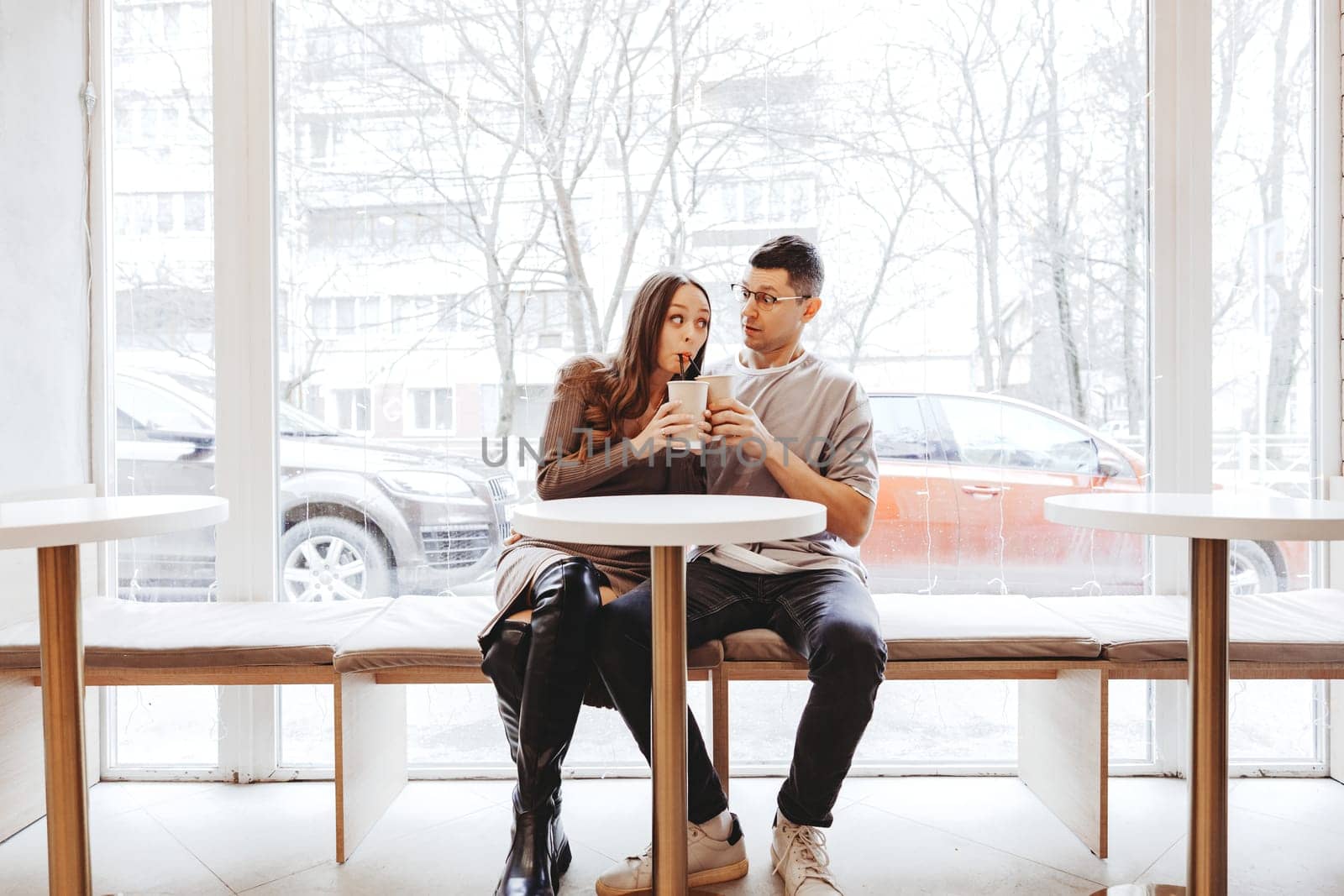 A couple shares playful moments while sipping coffee at a cafe. Frosted windows add a wintery backdrop to their cheerful interaction as they enjoy each other's company.