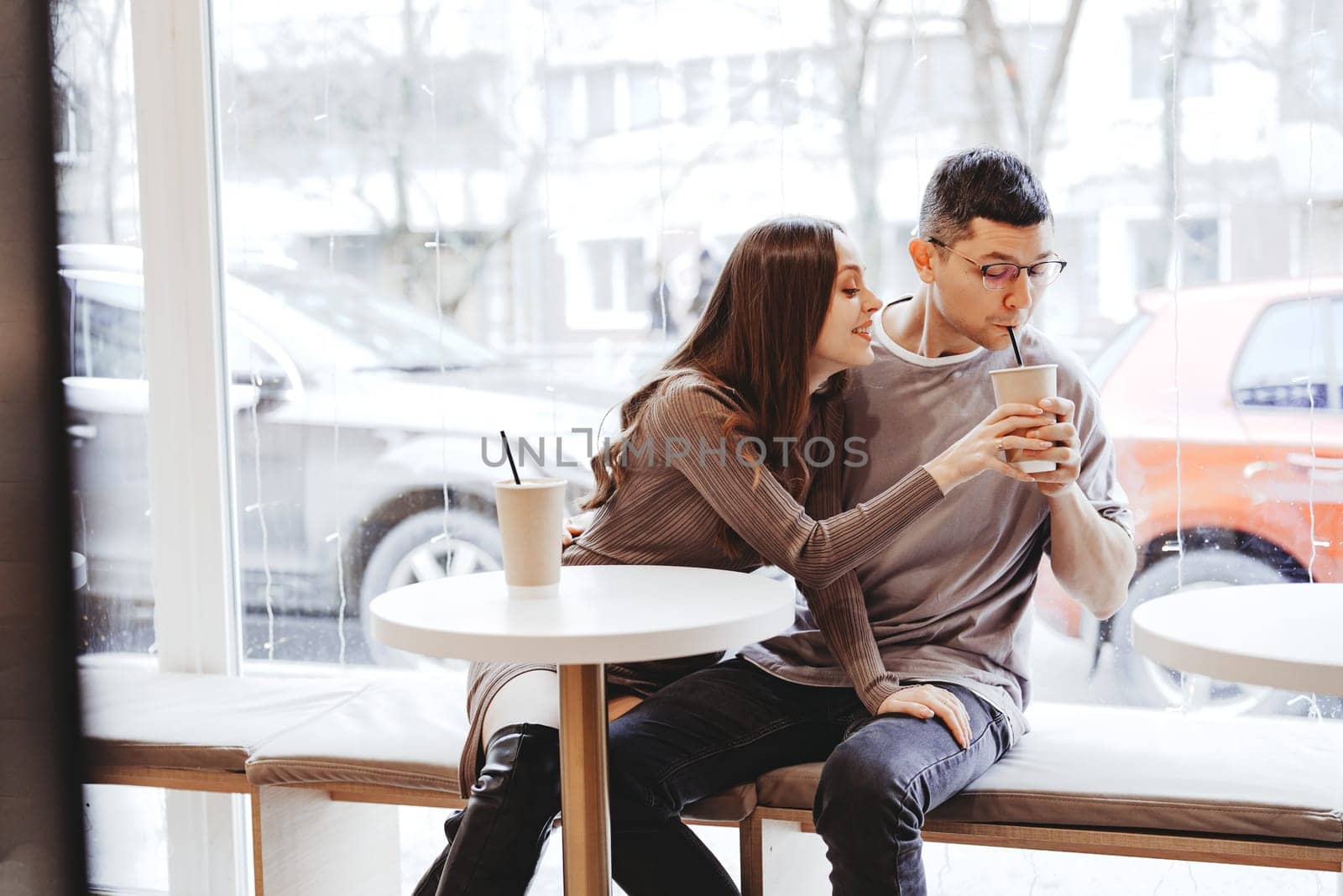 A young couple sits close together at a table in a cafe, sharing drinks and enjoying each other's company while snow falls outside.