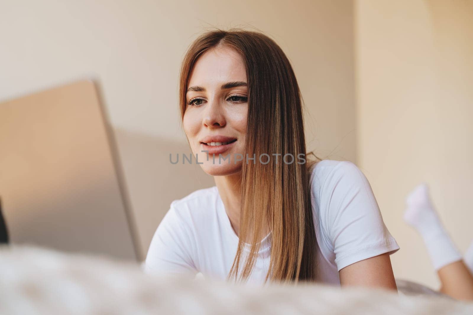 A young woman sits comfortably on a bed, focused on her laptop. Soft sunlight fills the room, creating a cozy atmosphere as she engages with her work.