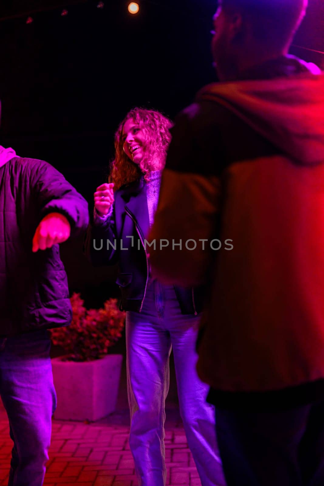 A group of friends is having fun dancing and chatting at an outdoor gathering. Colorful lights illuminate the space, creating a festive atmosphere as they move to music.
