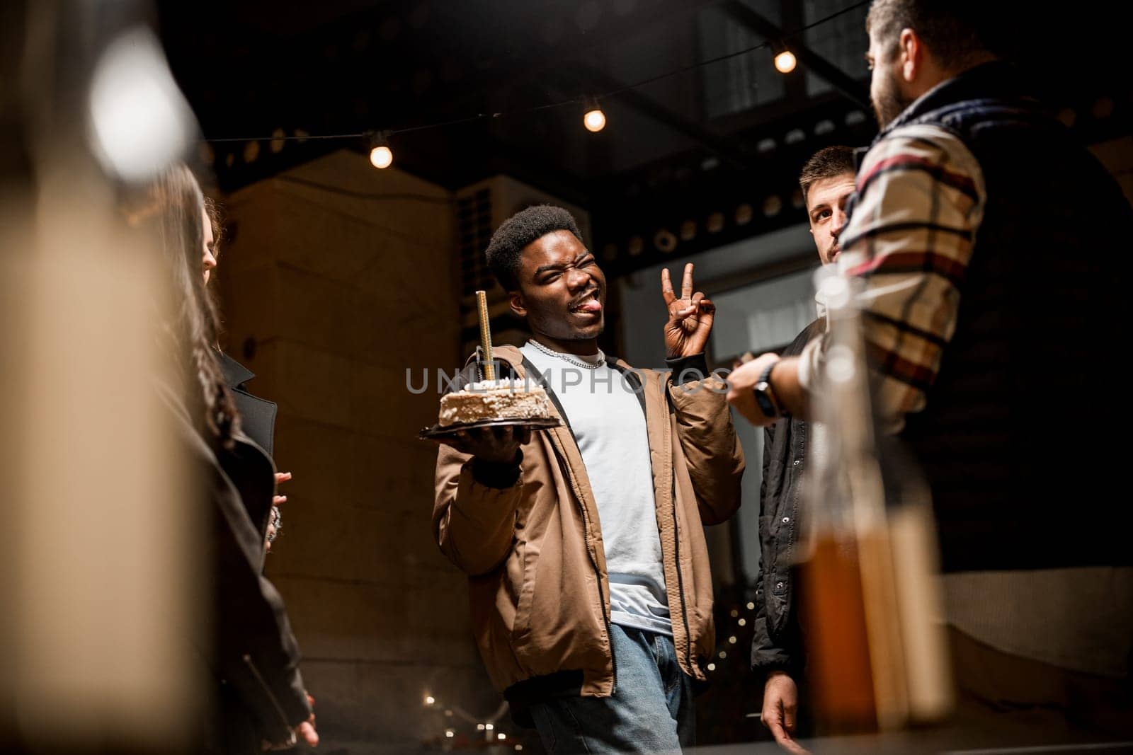 A group of friends gathers in a cozy setting, celebrating a birthday. One individual holds a cake, smiling and making a playful gesture. The atmosphere is festive and warm.