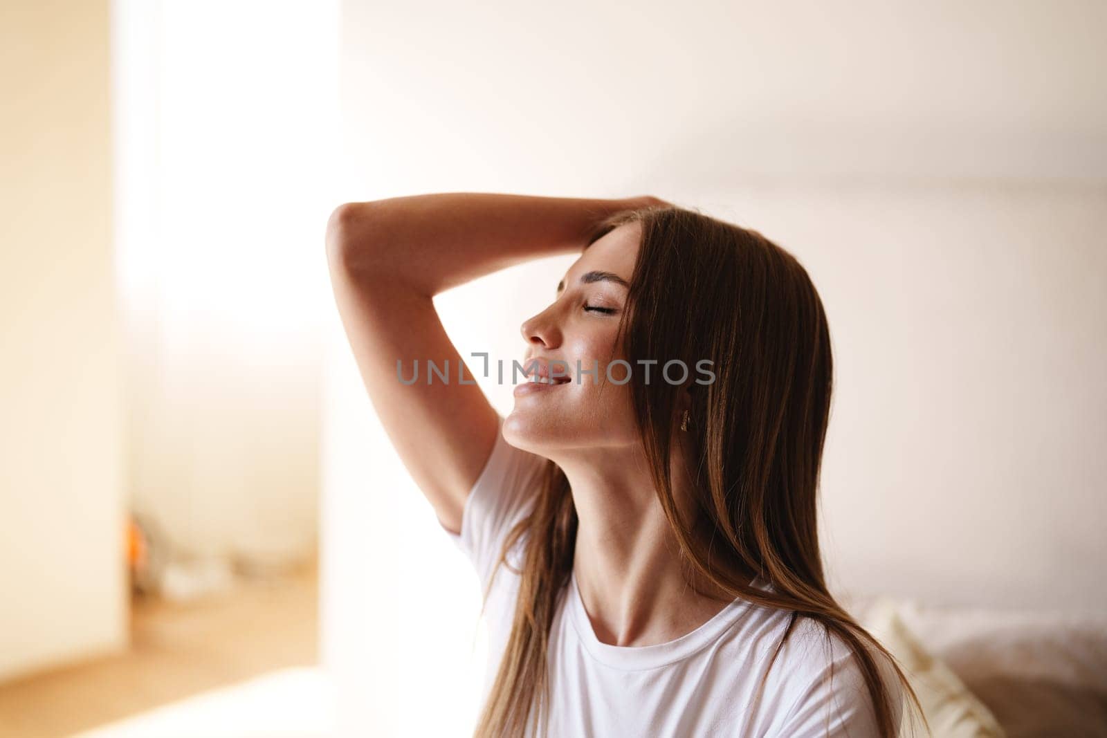 A young woman with long hair smiles gently while sitting in a bright, airy room. Sunlight filters through, creating a warm, peaceful atmosphere as she relaxes.