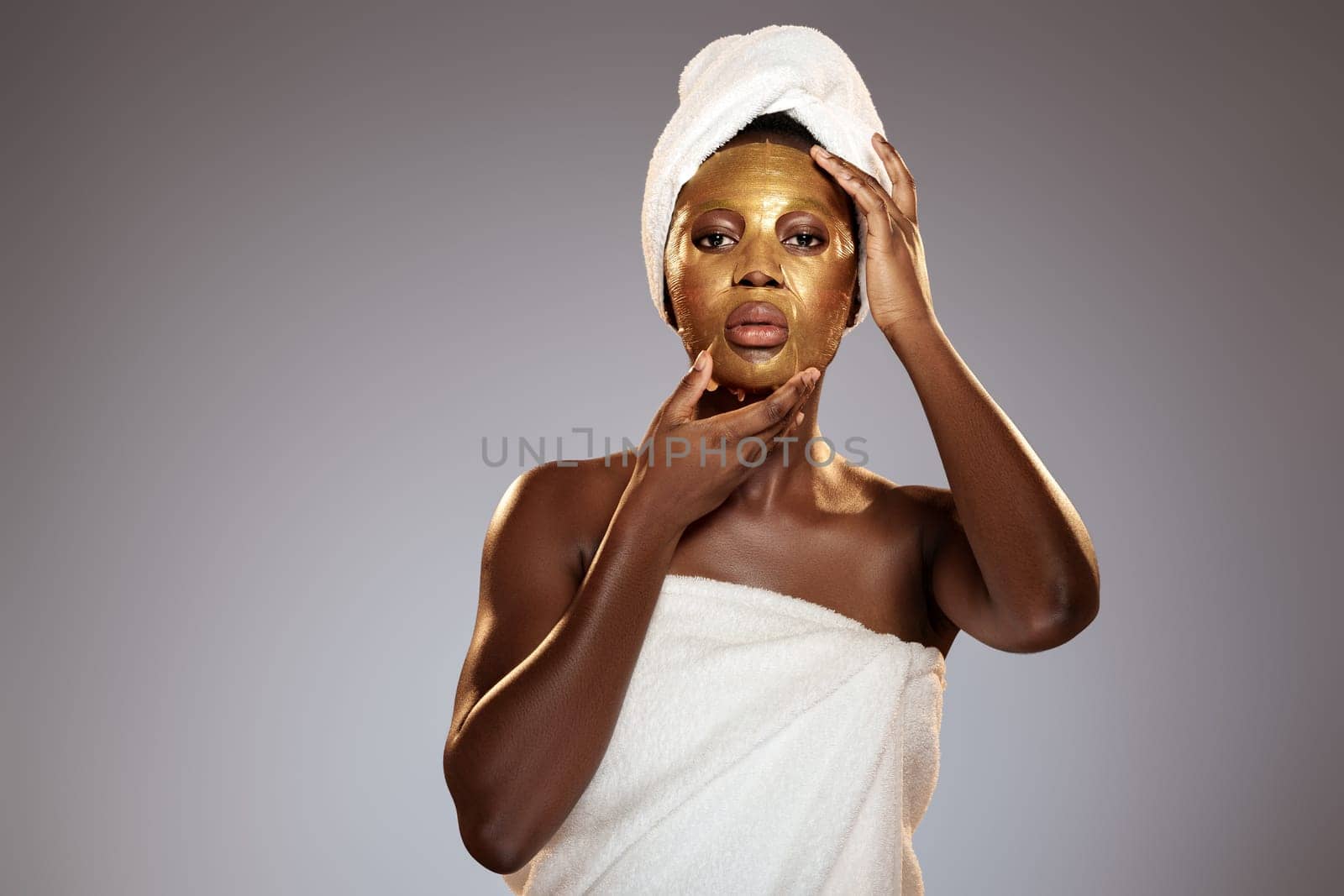 A woman stands in a calm, neutral-toned setting, applying a gold facial mask on her face. She is wrapped in a towel and has an additional towel on her head, embodying a self-care moment.
