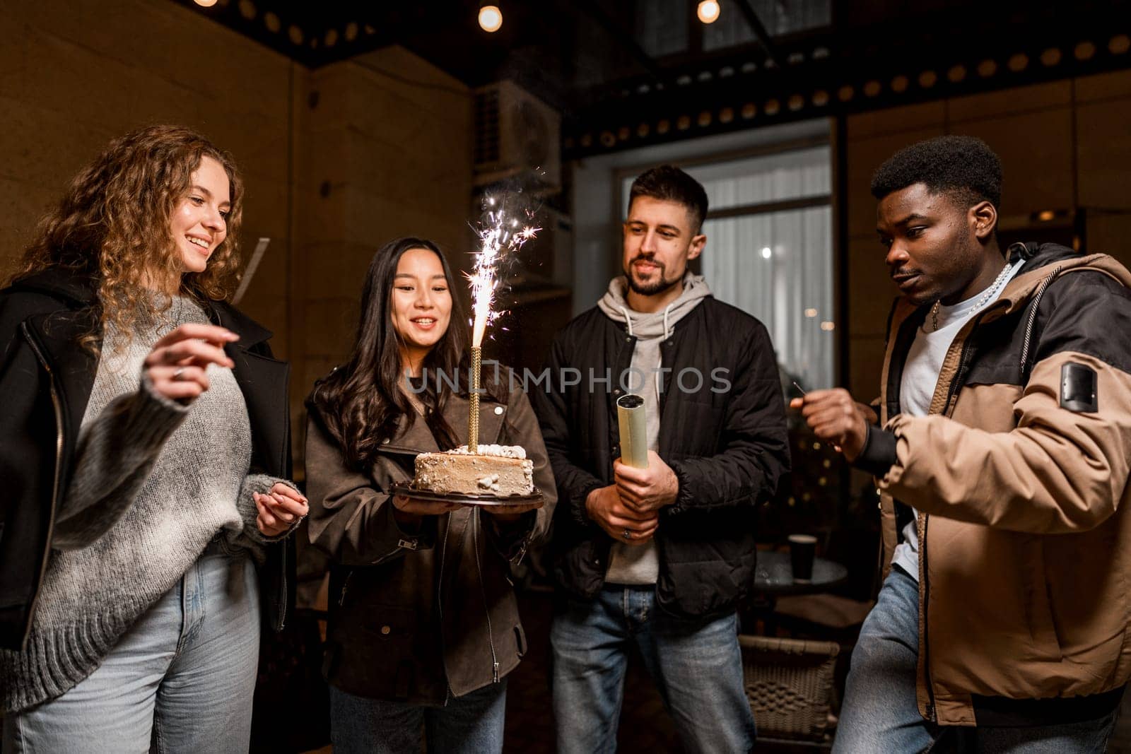 A group of four friends joyfully celebrate a birthday inside a cozy venue. One person holds a cake topped with sparklers as the others cheer and enjoy the moment together.