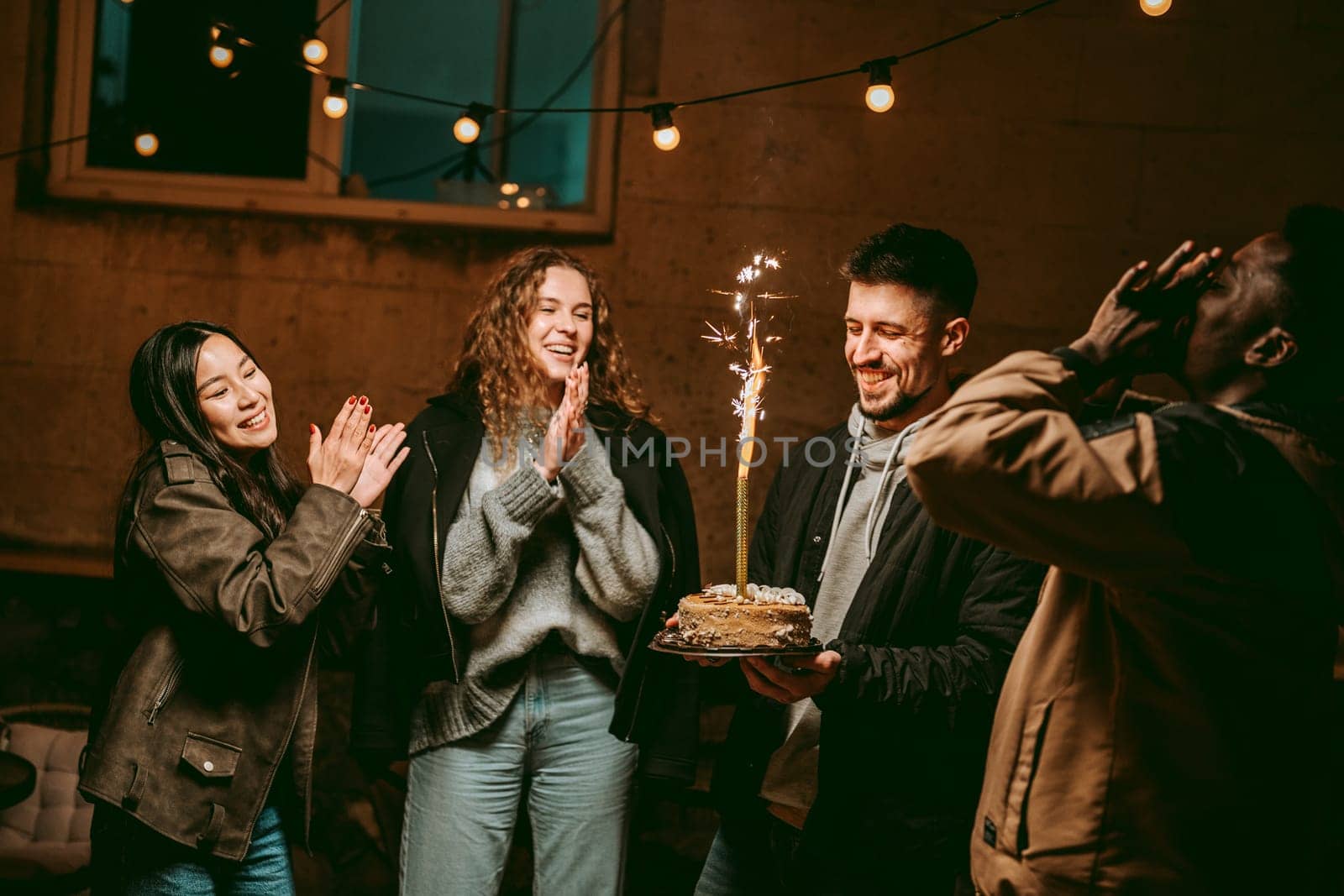 Four friends joyfully celebrate with a cake featuring sparklers on a cool evening. Laughter fills the air under warm string lights, creating a festive mood.