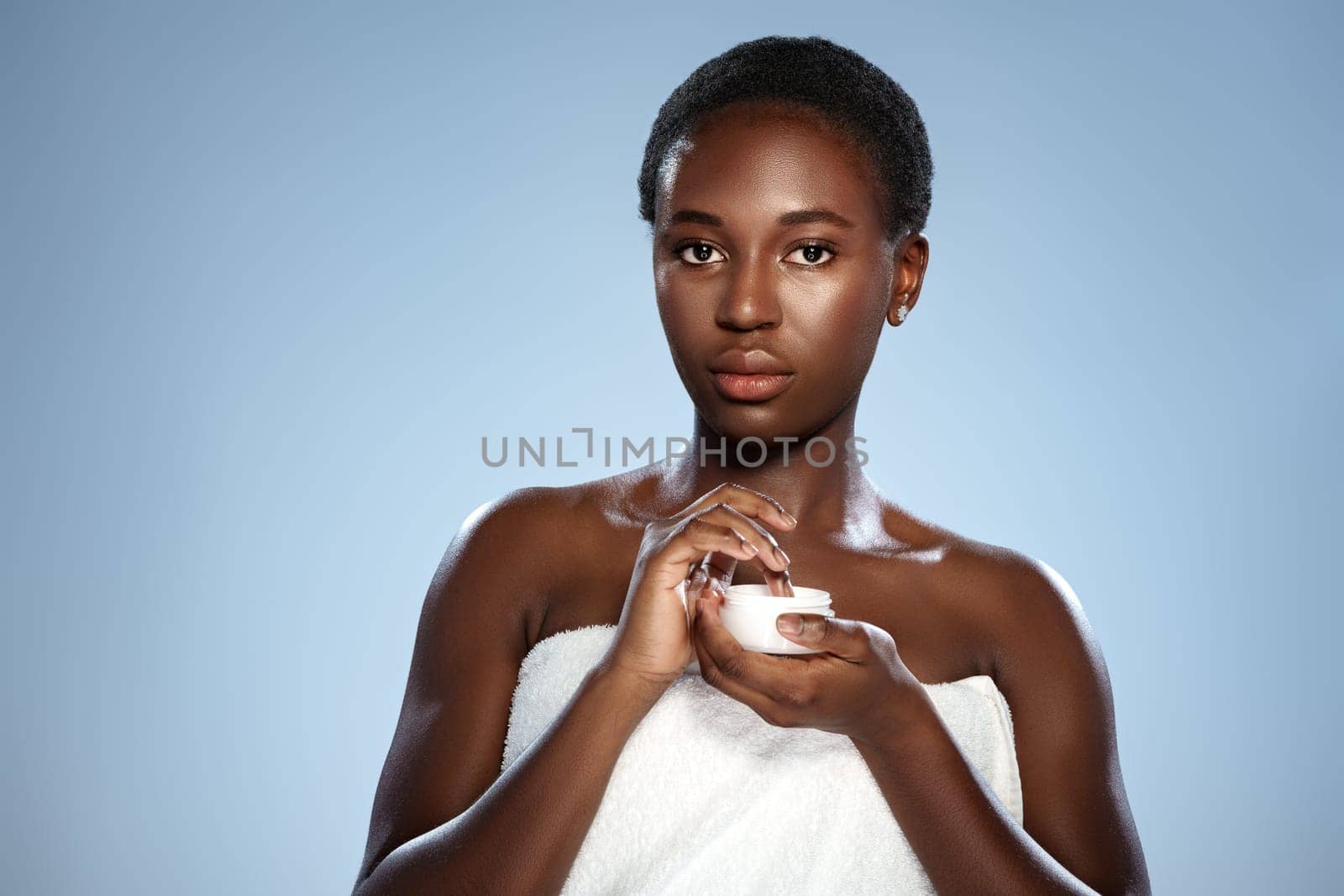 A woman stands with a towel wrapped around her, applying cream to her skin. The soft blue backdrop enhances the focus on her natural beauty and skincare routine, highlighting self-care.