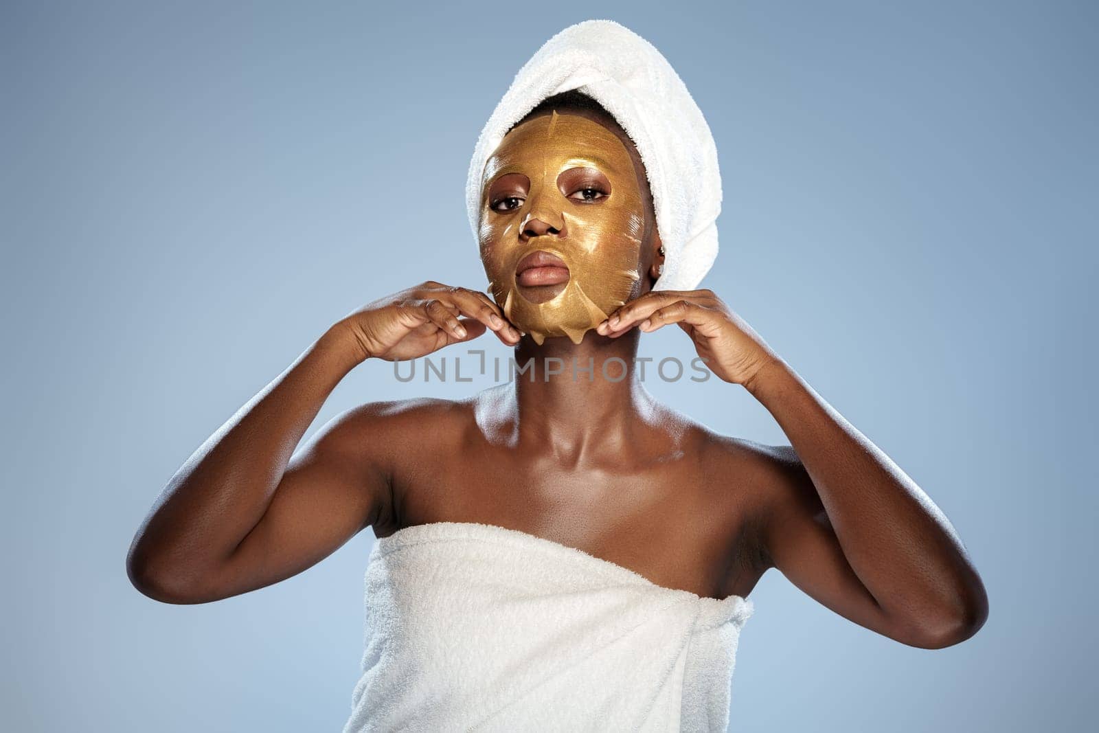A woman stands confidently, wearing a gold facial mask and a towel on her head. She is preparing for a skincare routine in a bright, calming environment, focusing on her beauty regimen.