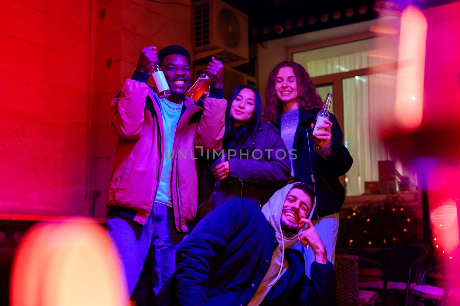 Group of four friends enjoying a night together, holding drinks and celebrating in a vibrant atmosphere filled with colorful lights and decorations.
