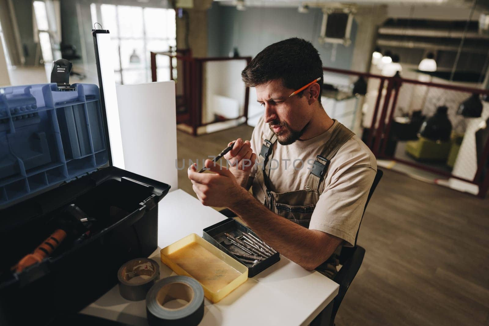 A skilled worker wearing overalls is carefully sorting tools in a workspace. The modern office features metal ductwork and clean, minimalist design, indicating ongoing repairs.