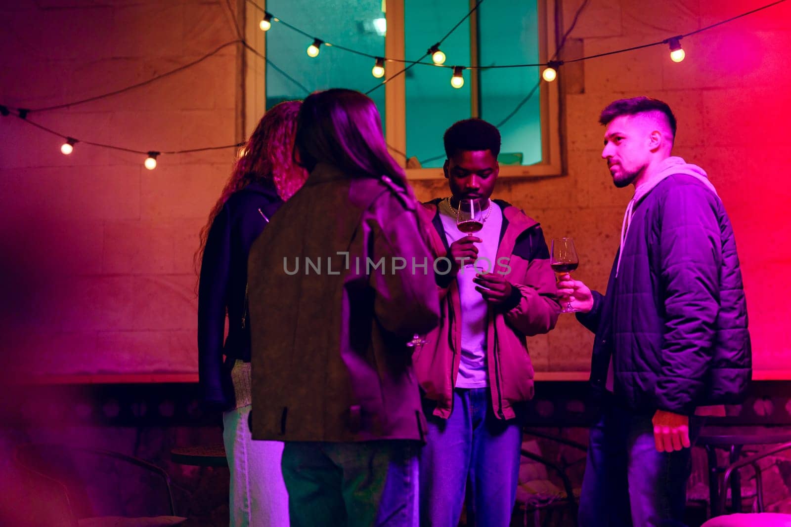 In a lively outdoor setting, a group of friends shares drinks and laughter under vibrant string lights, creating a festive evening ambiance.