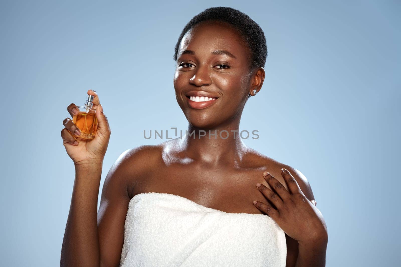 A woman with short hair is smiling while holding a perfume bottle in a bright studio. She is wearing a white towel and exudes confidence and joy. The soft blue backdrop enhances the mood.
