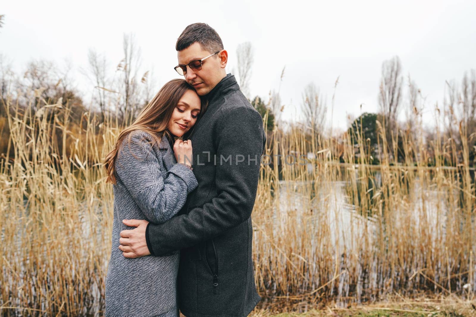 A couple stands closely together by a quiet body of water, surrounded by tall reeds. The woman rests her head on the man's shoulder, both wearing warm coats as they enjoy a calm autumn day.