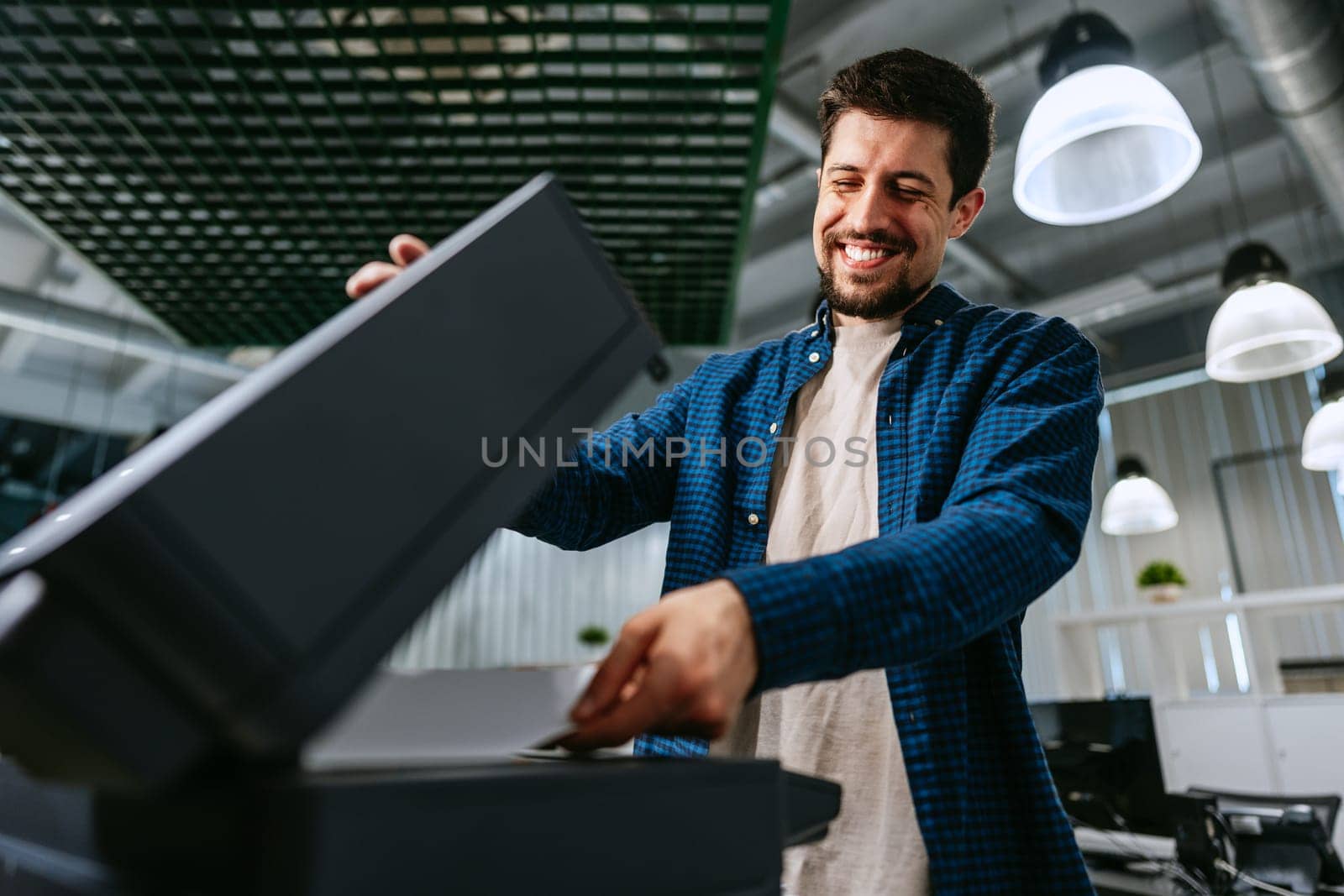 A man smiles while interacting with a printer in a contemporary office space filled with overhead lights and computers. He appears focused on his task.