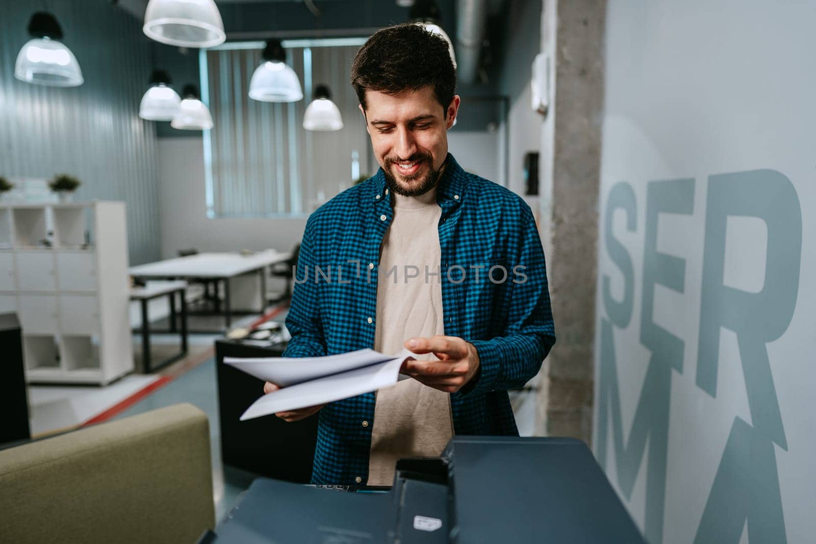 A young man stands in a contemporary office, smiling as he reviews printed papers from a printer. Natural light fills the workspace, creating an inviting atmosphere for productivity.