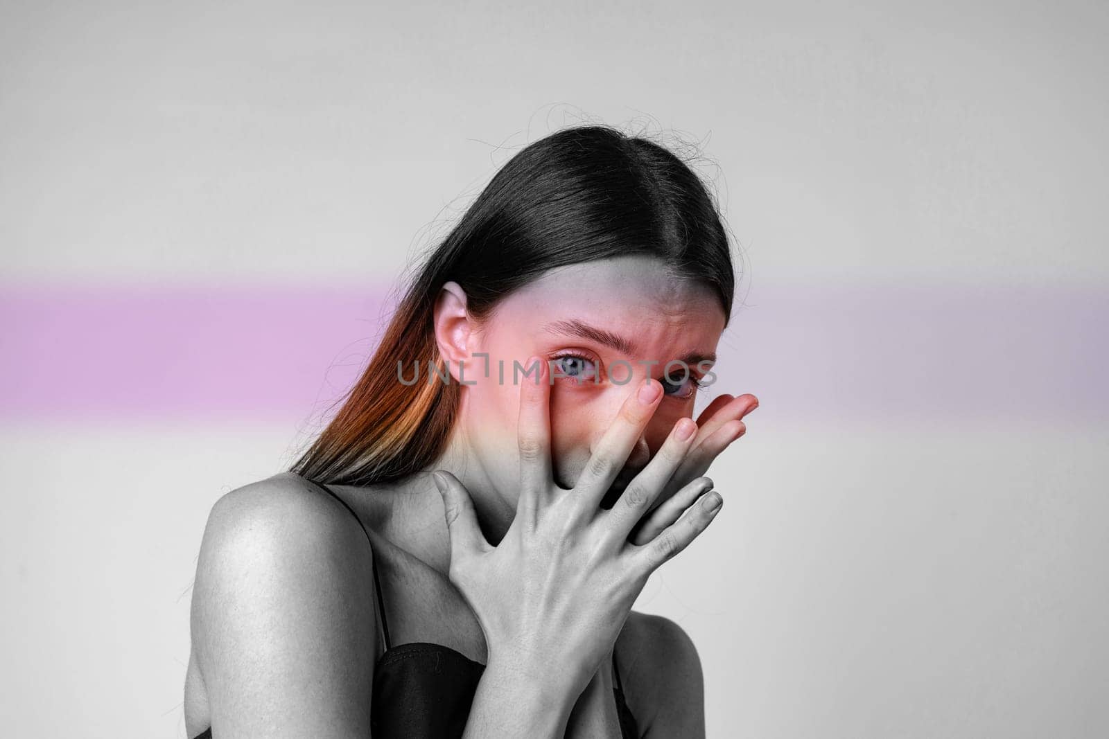 A woman stands in a minimalist setting, daylight illuminating her features. She expresses deep emotion with her hands covering her face, revealing striking blue eyes.