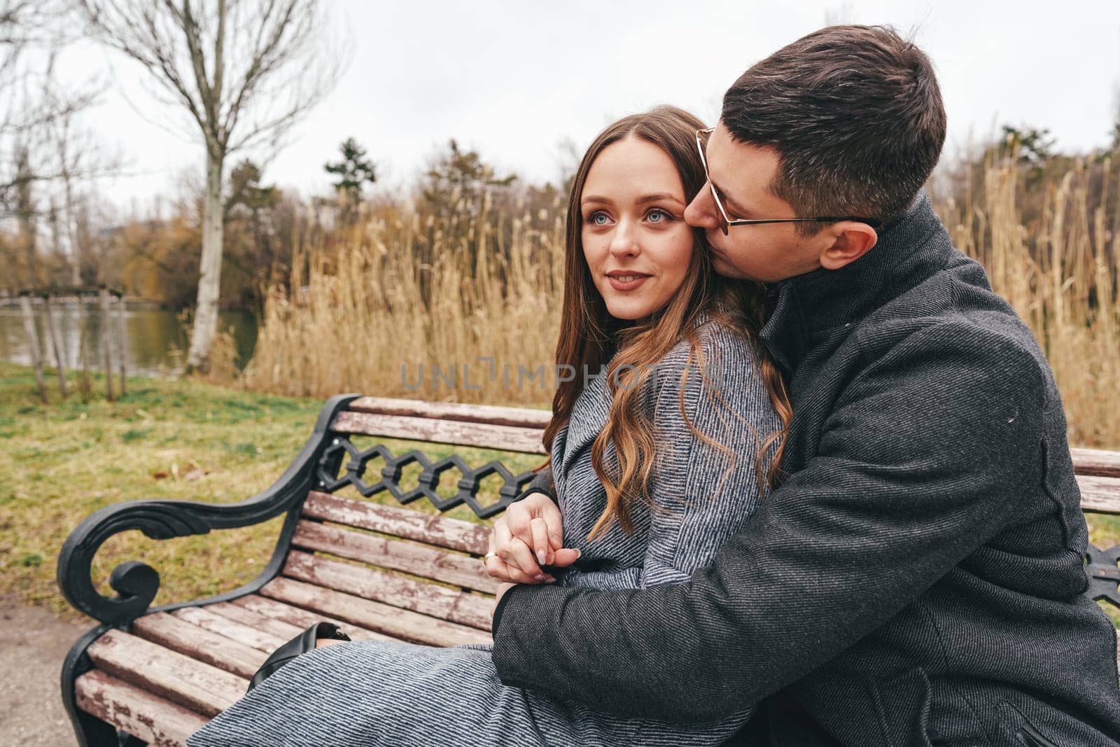 A young couple sits closely on a park bench, sharing a warm moment. They are surrounded by tall grass and trees in a serene outdoor setting during autumn.