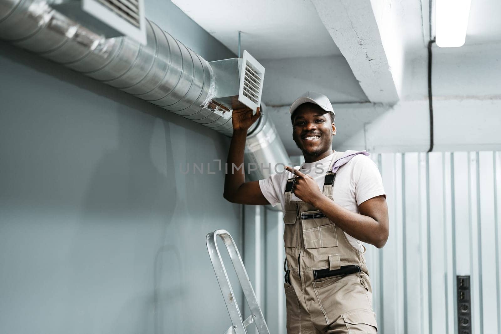 A professional worker wearing a cap and overalls stands next to a newly installed air vent in a workshop. The person is smiling and pointing, showcasing expertise.