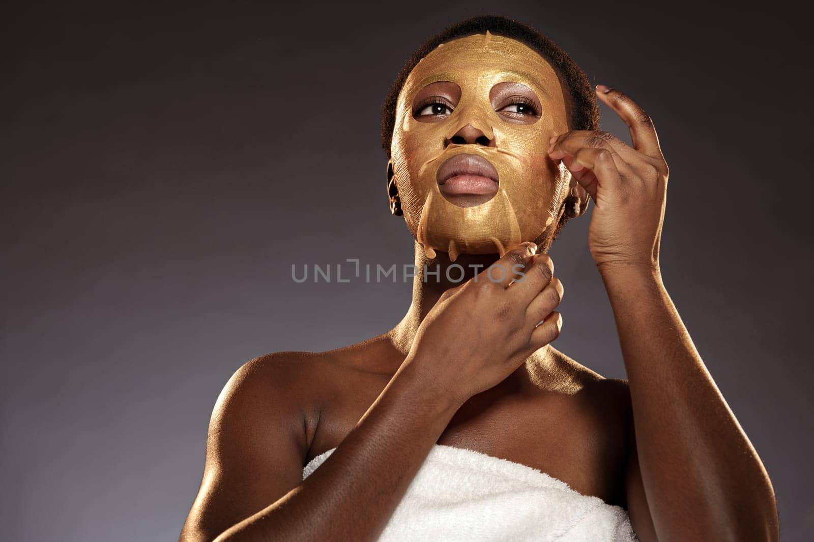 A woman is gently removing a gold facial mask while wrapped in a towel. The setting has a calm, soothing atmosphere, ideal for skincare and relaxation.