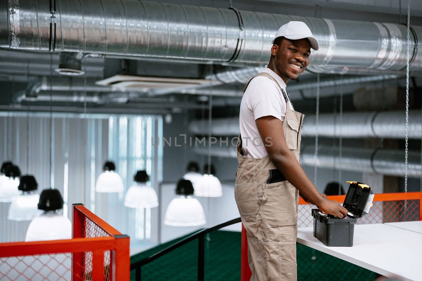 A young male worker smiles while using a tool on a table in a contemporary office environment filled with pendant lights. Bright ambiance enhances his cheerful demeanor.