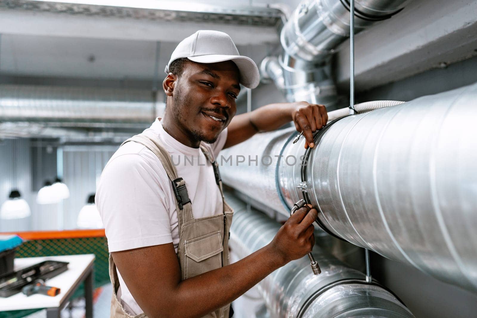A technician wearing a cap and work overalls tightens a fitting on a large metal pipe in an industrial setting, showcasing expertise and focus on the task at hand.