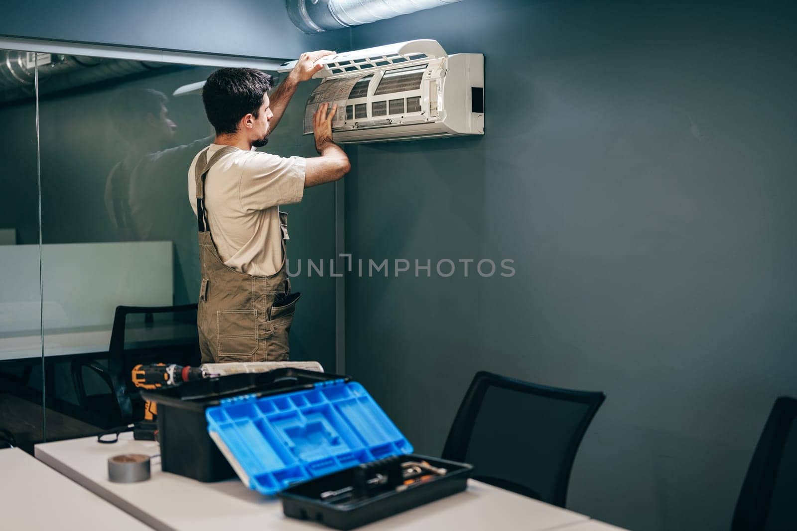 A skilled technician adjusts an air conditioning unit mounted on a wall in a contemporary office space. Tools and equipment are visible on the table nearby.