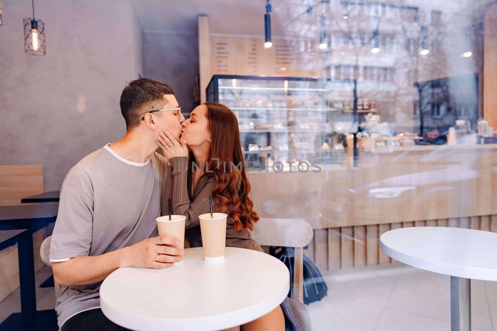 A young couple enjoys their time at a cafe, sitting at a small table with iced drinks. They share a kiss, creating a romantic atmosphere in the cozy setting.