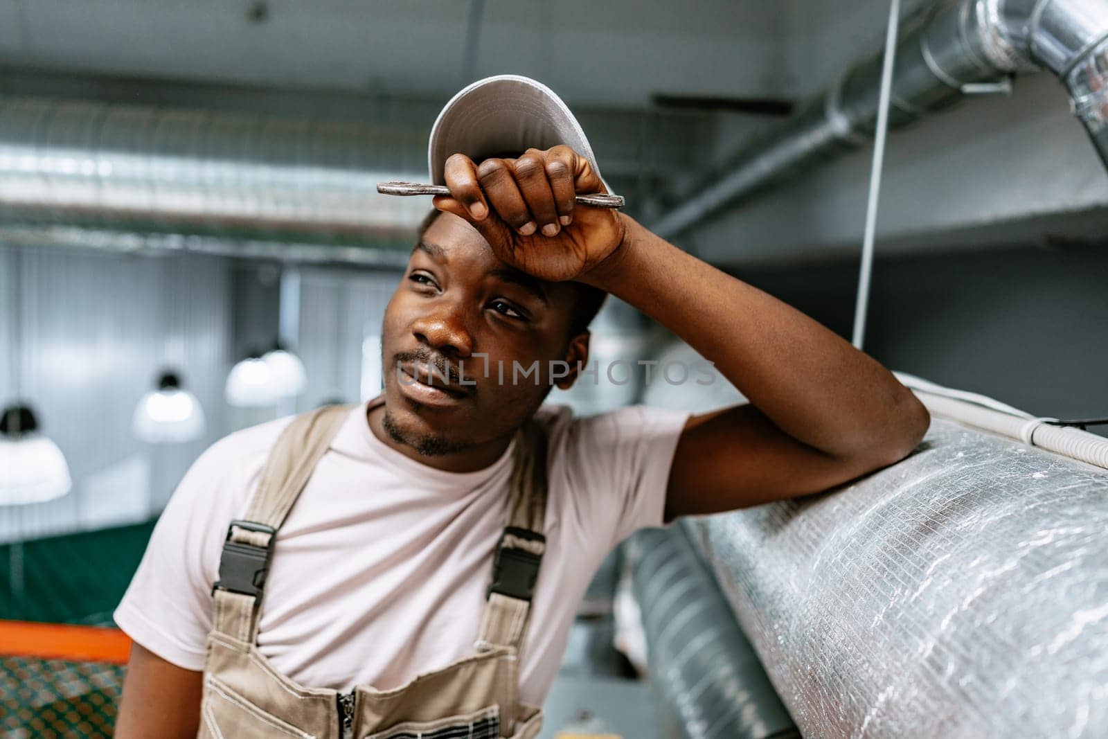 A young man stands with his hand on his forehead, wearing a cap and overalls. He appears thoughtful in a well-lit industrial workspace with metal ductwork overhead.