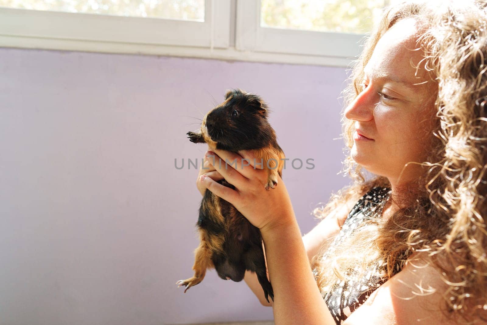 A woman gently holds a guinea pig with a smile in a bright, sunlit room. The atmosphere is warm and inviting, showcasing their special connection.