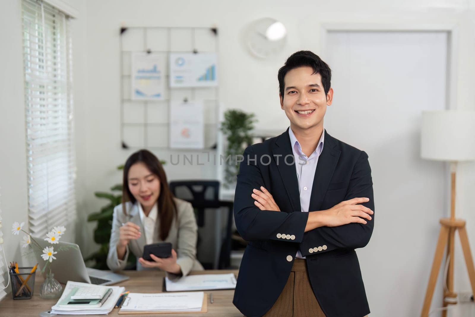 A confident businessman stands with arms crossed in a bright office, showcasing professionalism and collaboration.