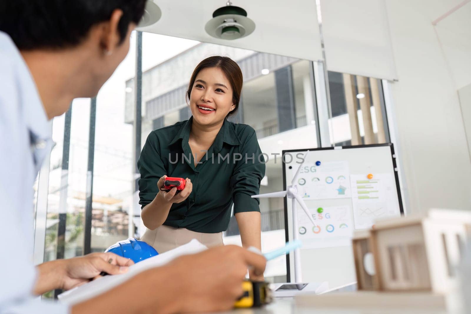 A smiling female architect engages with a colleague in a modern office, discussing project details and sustainability.