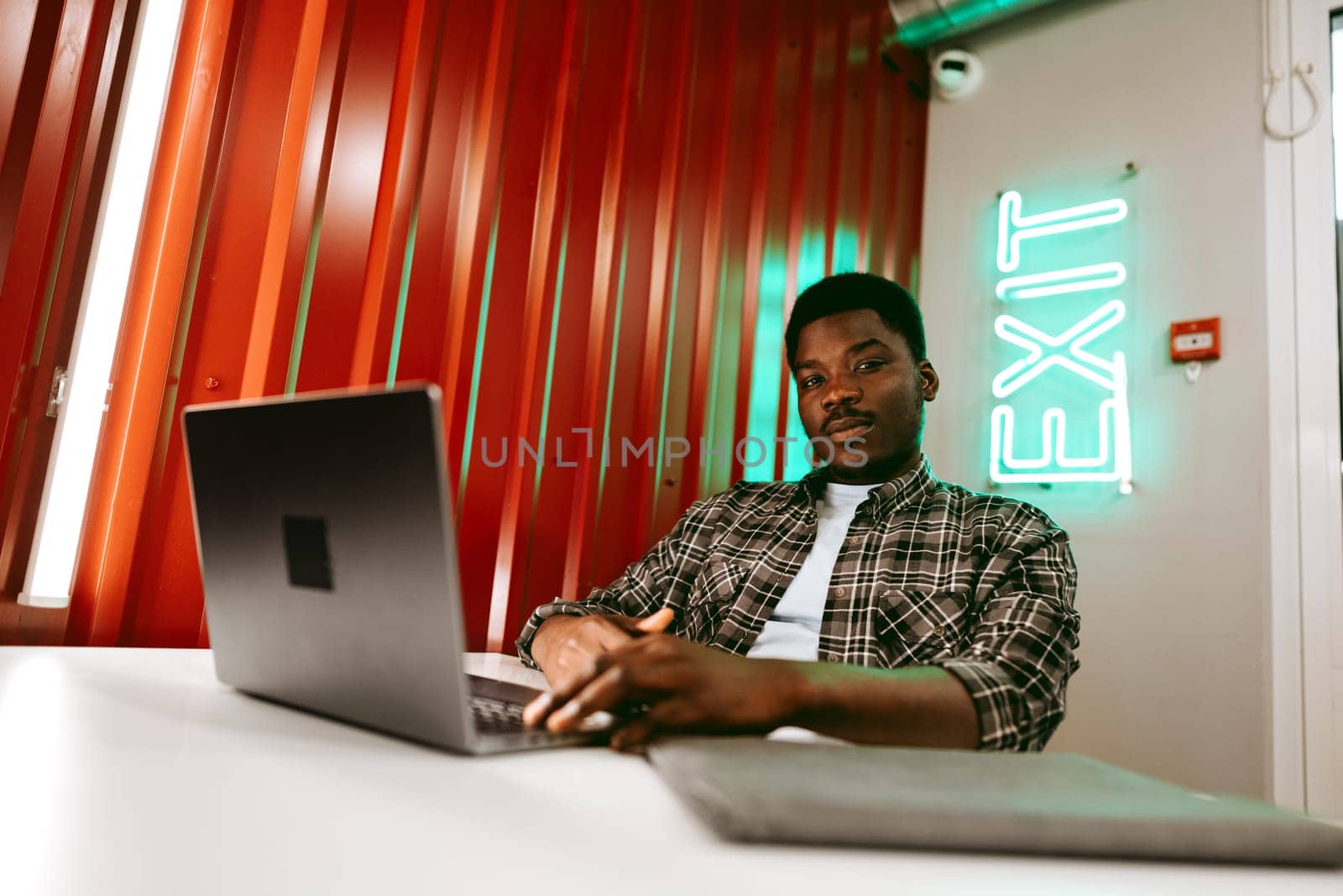 A young man sits at a table in a contemporary workspace, focused on his laptop. The striking red walls and neon exit sign create an energetic atmosphere.