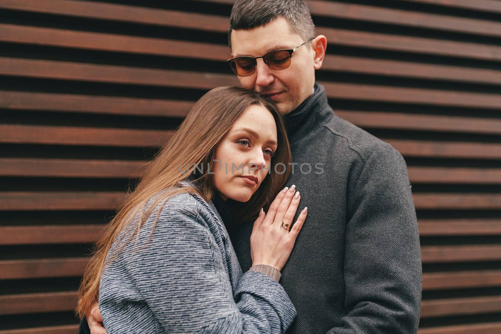A couple stands close together, sharing an affectionate moment. The woman leans into the man, who wears sunglasses, creating an intimate atmosphere amidst a modern urban backdrop.