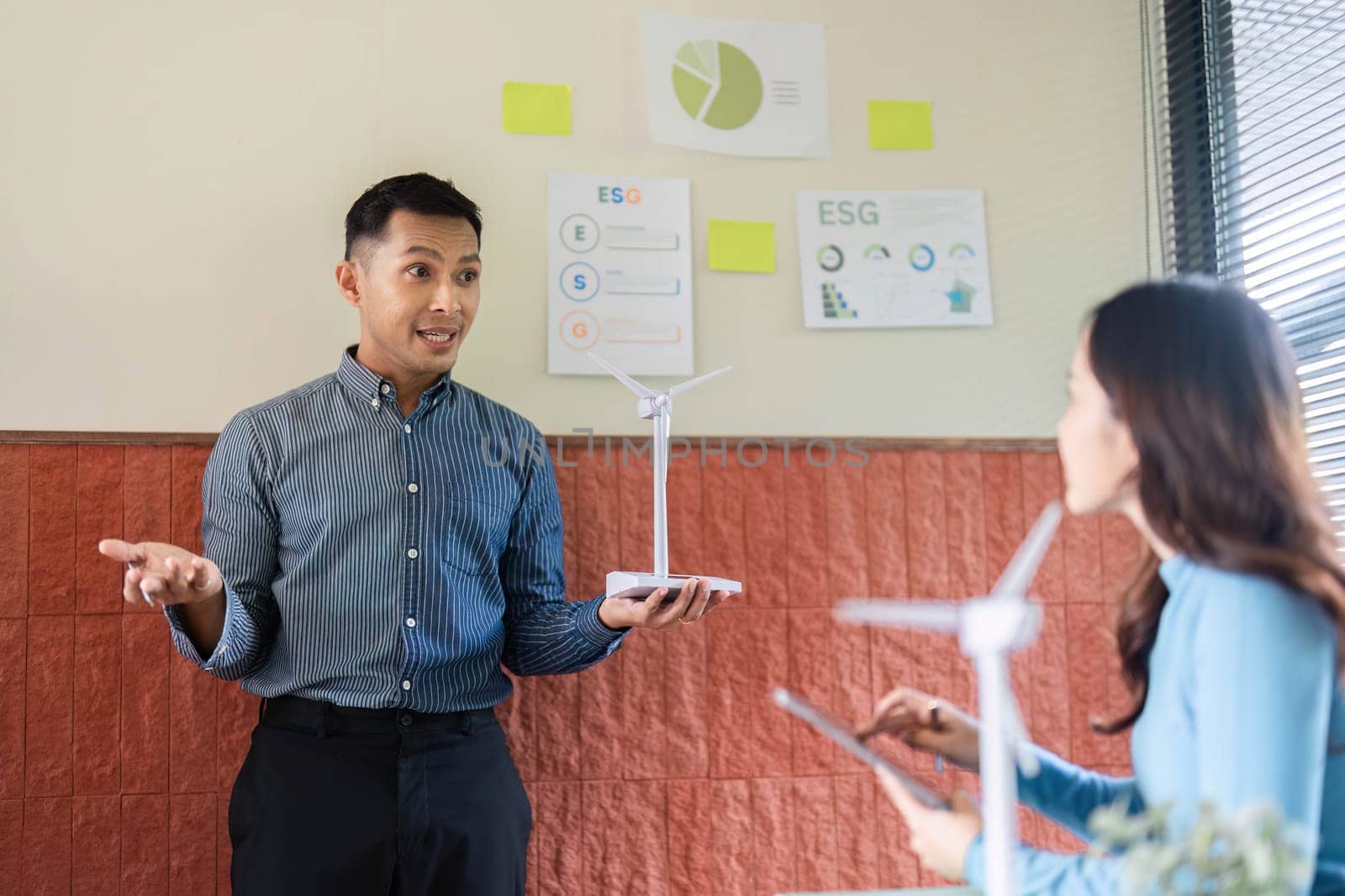 A professional presents a wind turbine model to a colleague, discussing the importance of ESG in renewable energy projects.