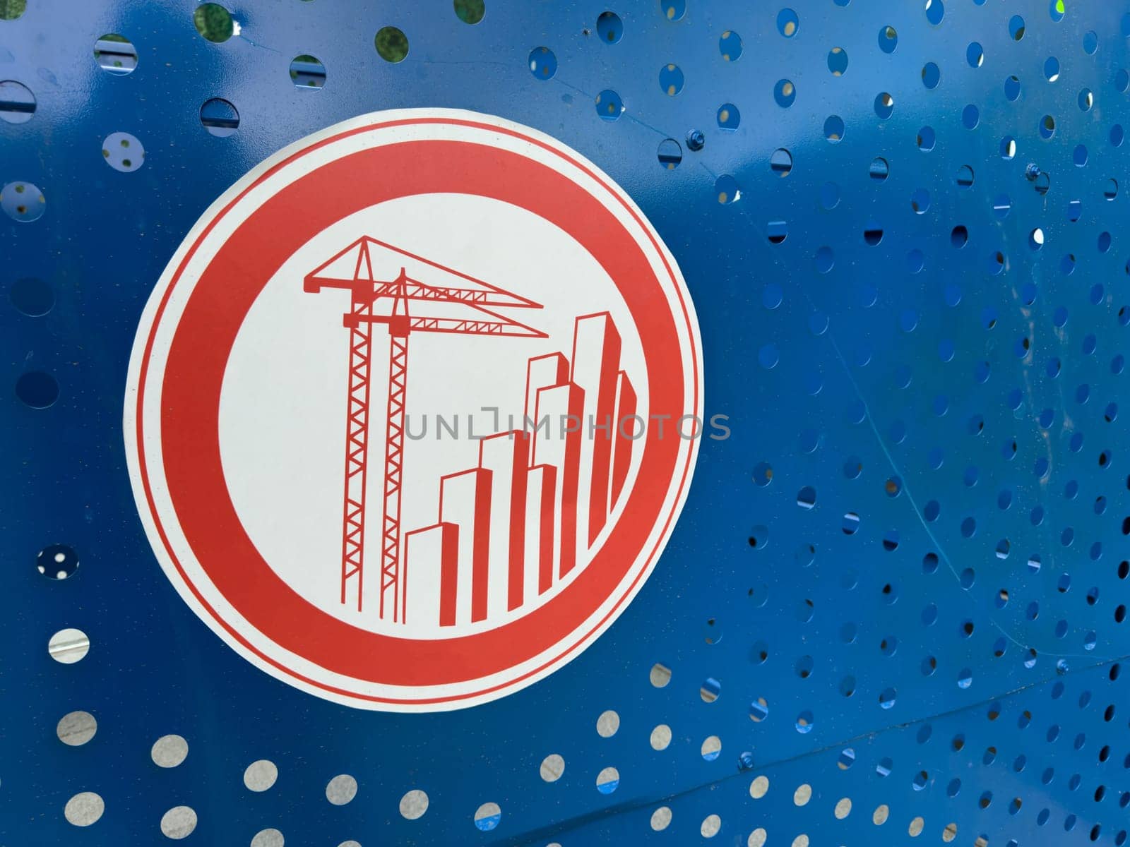 Red construction themed warning sign with cranes and skyscrapers printed on perforated blue metal fencing, symbolizing active development zone and modern urban growth initiatives