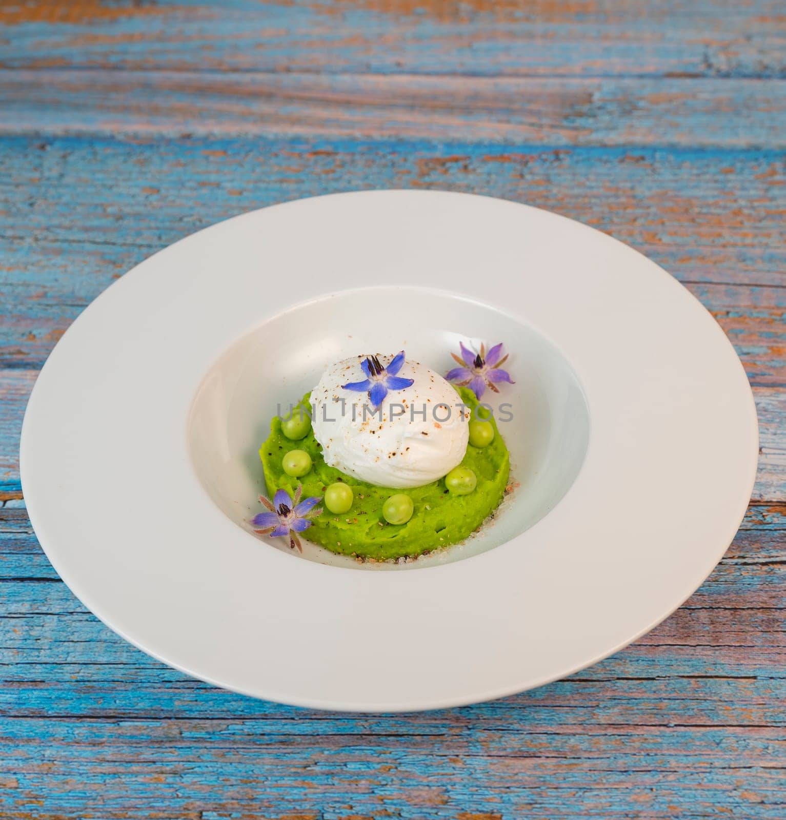 A fresh pea puree is beautifully plated, featuring a poached egg at the center and adorned with borage flowers. The dish showcases vibrant colors and textures, perfect for any spring menu.
