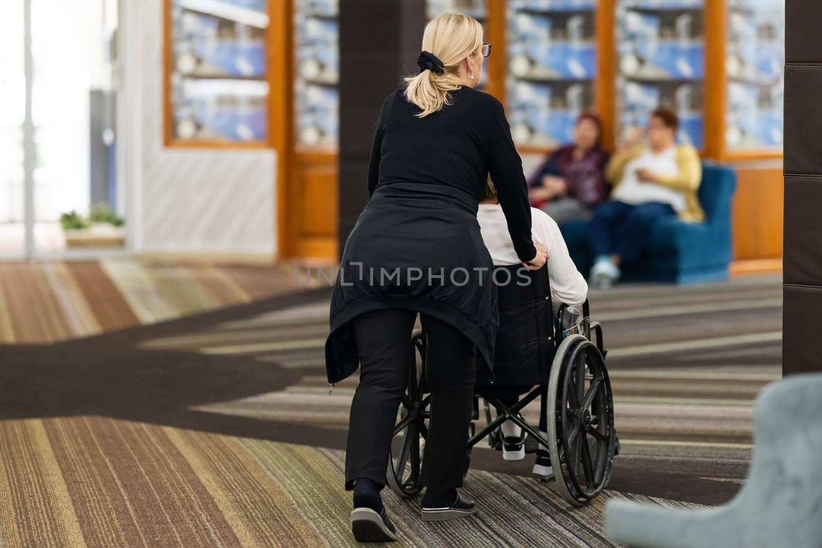 Rear view of a female caregiver pushing an elderly person in a wheelchair through a hallway.