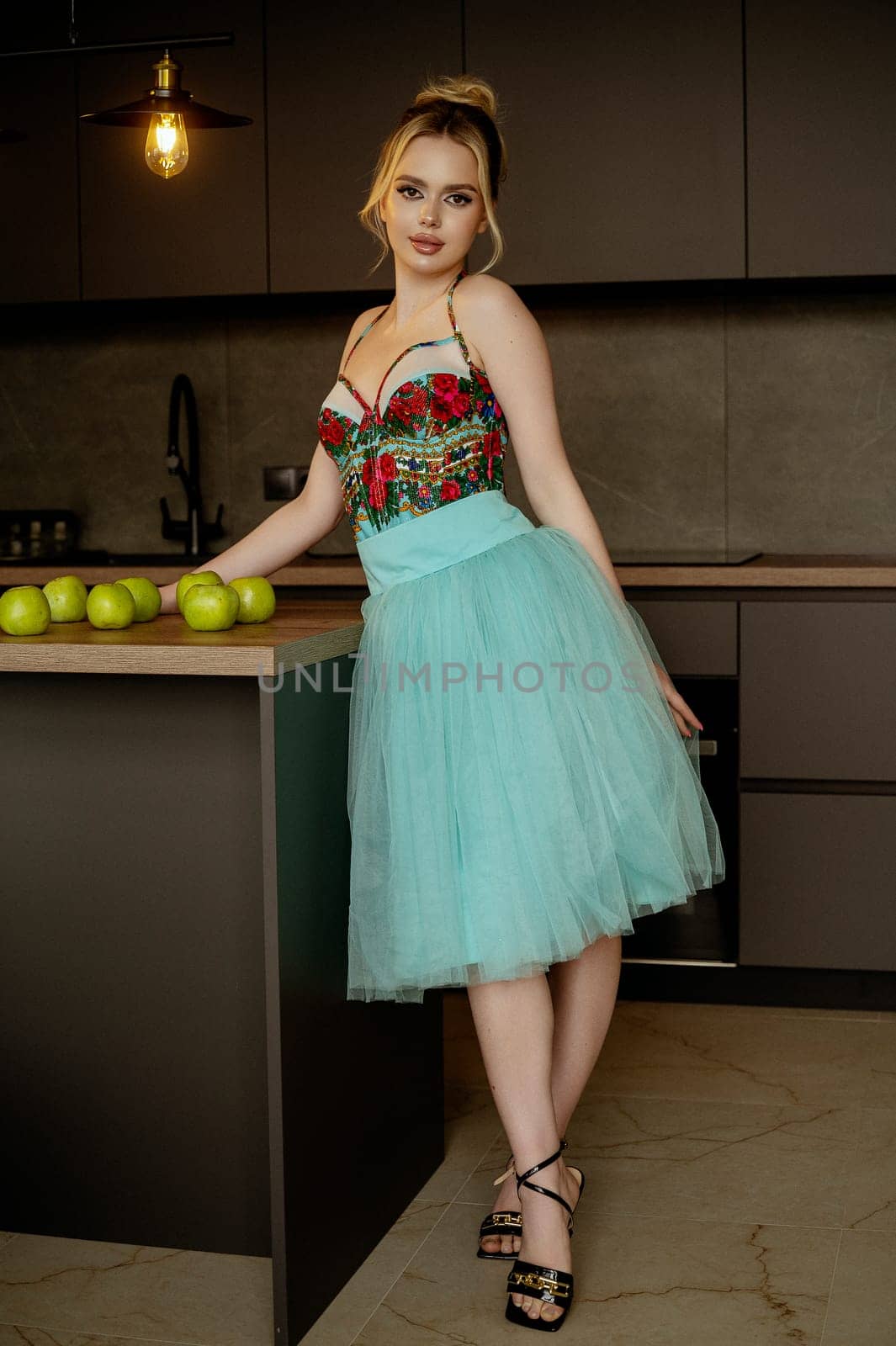 Elegant woman in turquoise skirt and corset gracefully poses in dark minimalist interior. demonstrating style, happiness and leisure. Modern kitchen, green apples on table, looking at camera