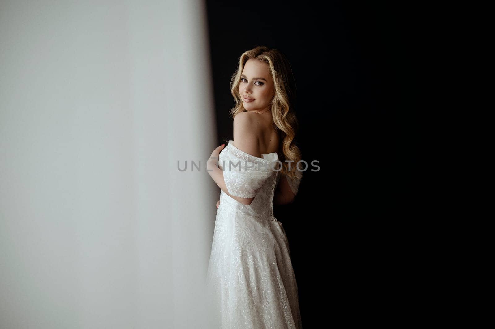 Elegant bridal gown showcased in a minimalist setting with a model in a chic pose during an indoor shoot. image of a blonde bride against a dark interior. smiles happily and looks at the camera