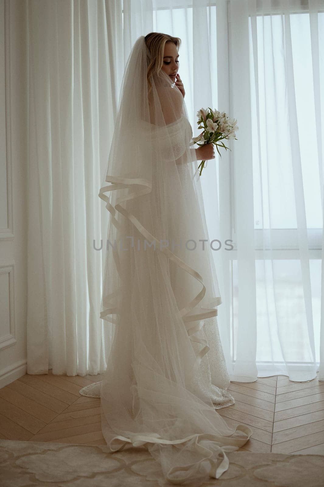Bride with luxurious long vulya and elegant dress poses against the background of the window. huge window with silhouette of bride with bouquet of lilies. Bride waiting for groom.