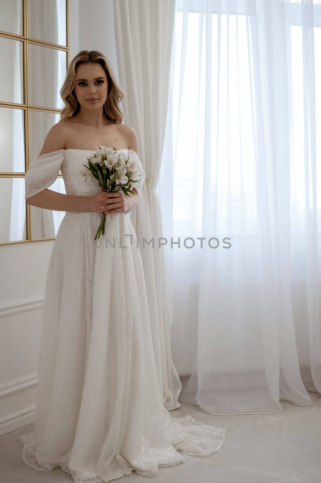 Bride with a bouquet of white flowers posing by the window. Blonde in a flowing wedding dress, radiates grace and beauty. In a light minimalist interior with a window and a mirror
