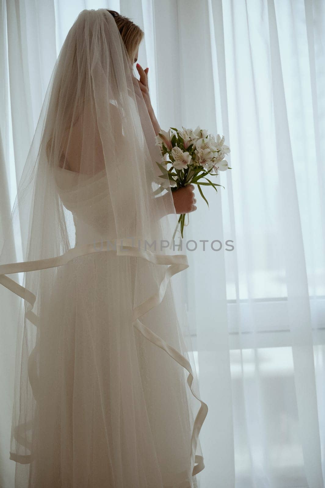Bride with luxurious long vulya and elegant dress posing against the window. huge window with silhouette of bride with bouquet of lilies. Bride waiting for groom and wiping away tears